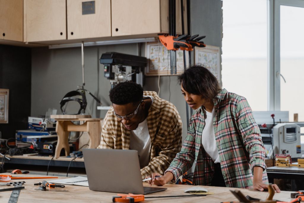A man and woman collaborating on a project using a laptop in a woodworking workshop.