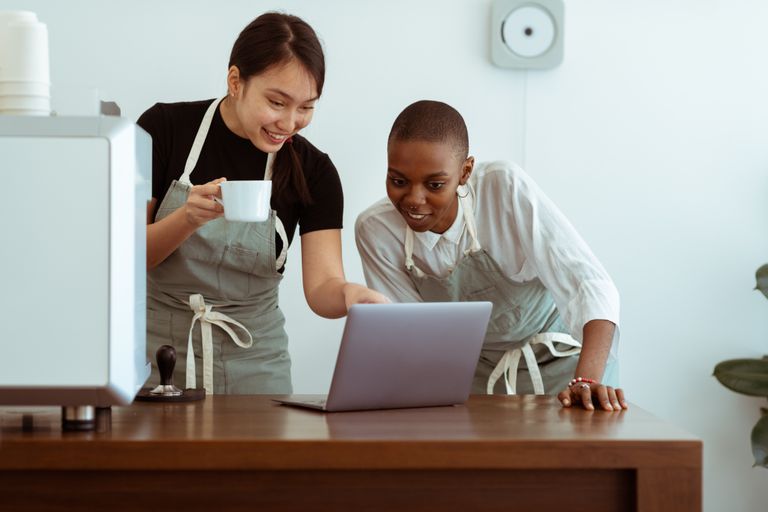 Positive smiling young multiracial waitresses wearing aprons gathering near table with cup of hor drink and surfing modern netbook in light kitchen
