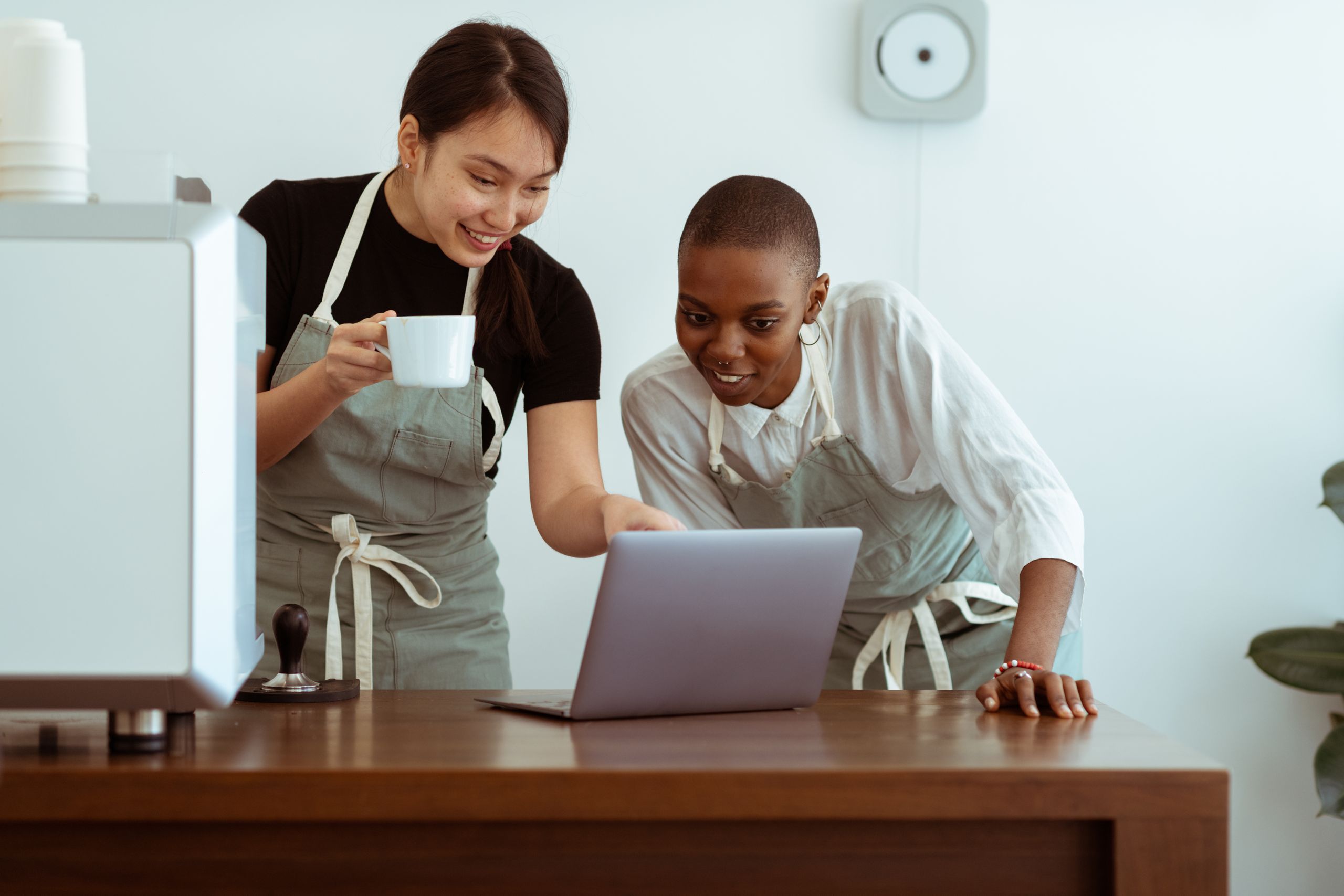 Positive smiling young multiracial waitresses wearing aprons gathering near table with cup of hor drink and surfing modern netbook in light kitchen